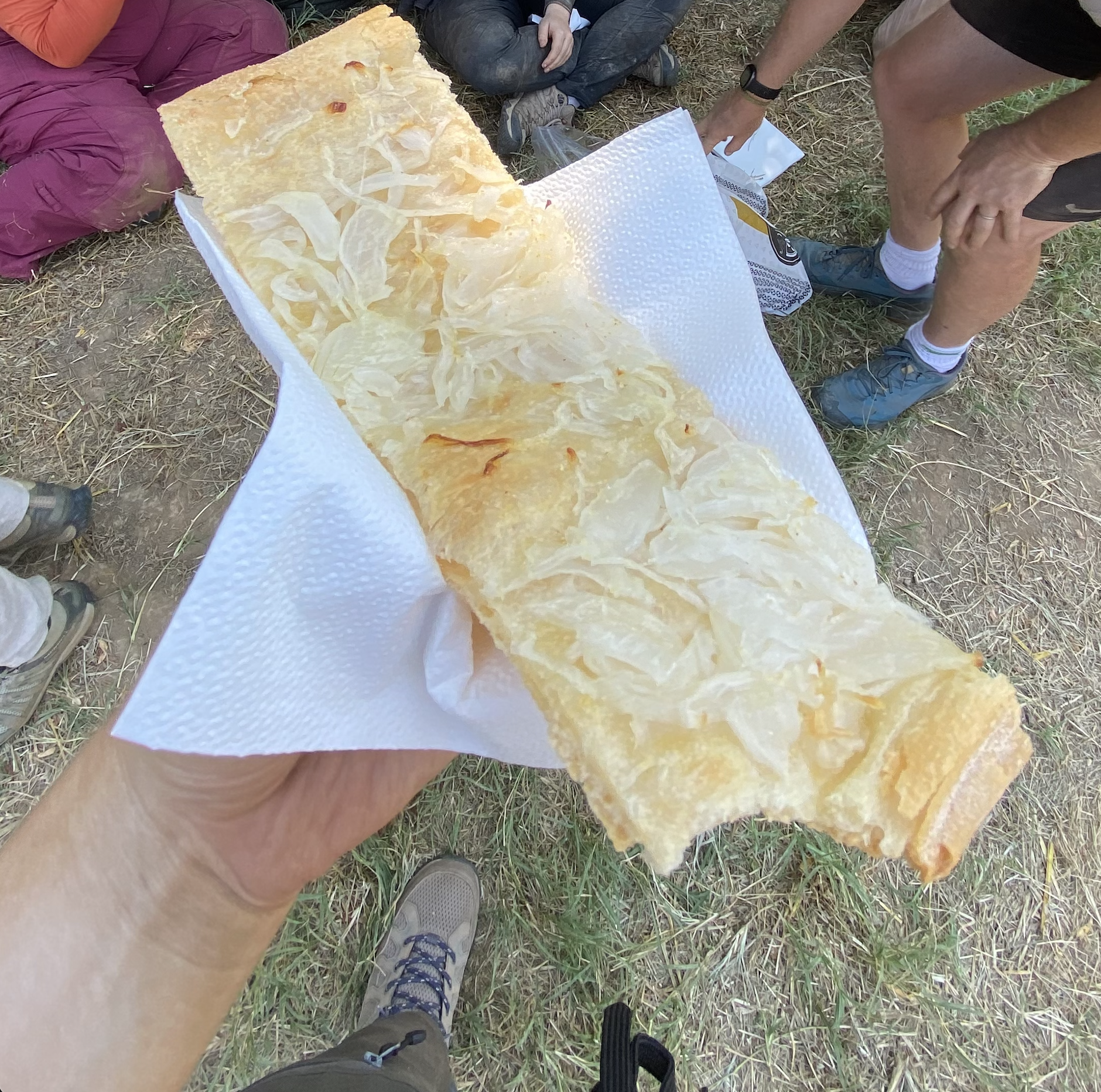 thin, flat bread covered in onions held in a hand with people sitting cross-legged in the dirt in the background